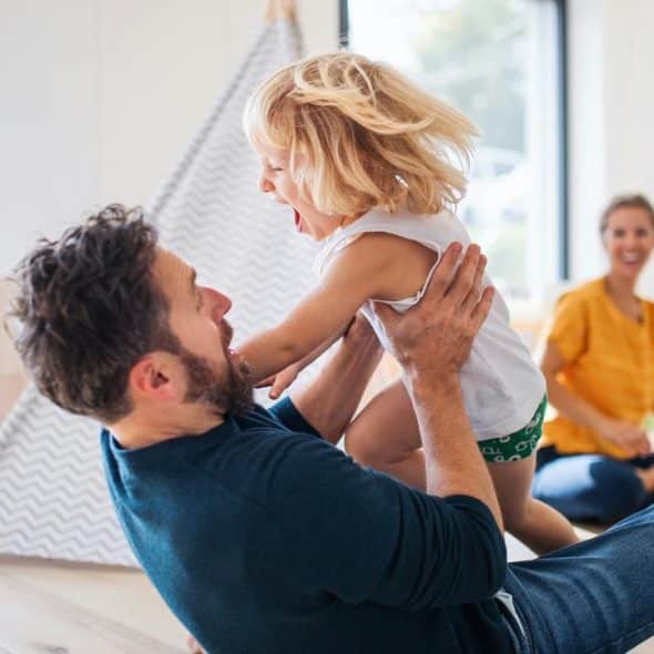 A father lifting his laughing toddler as the mother smiles behind.