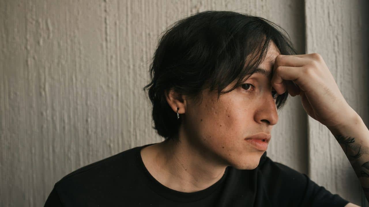 A somber person with dark hair looks to the side, with a hand on their forehead.