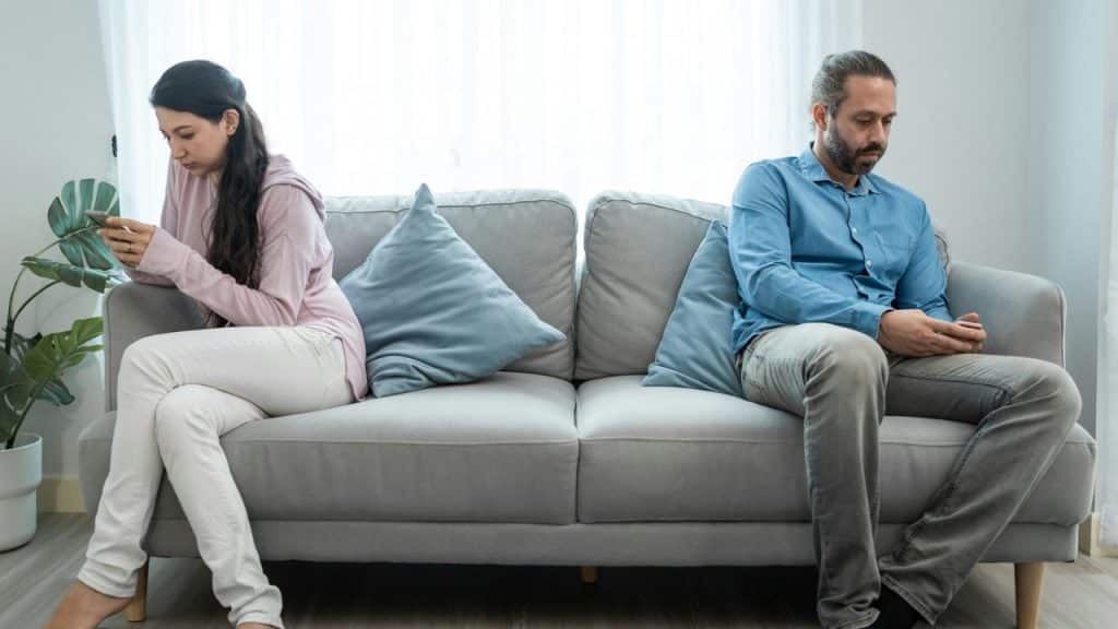 A man and woman sit separately on a sofa, both looking at their phones.