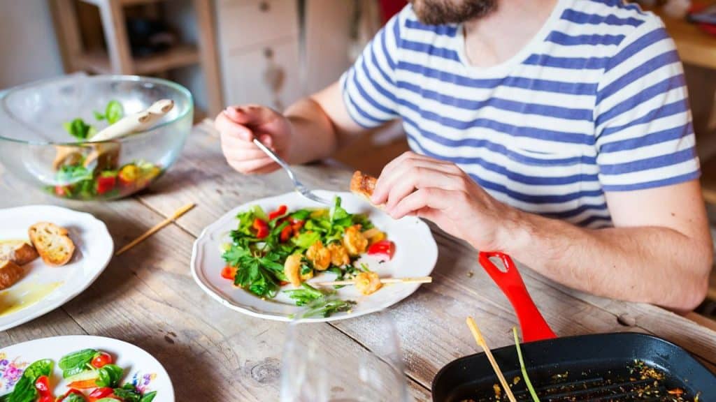 A man in a striped shirt eats a plate of salad with shrimp, sitting at a wooden table.