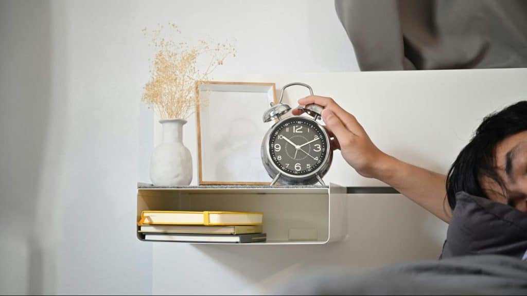 A hand reaching for an alarm clock on a bedside shelf.