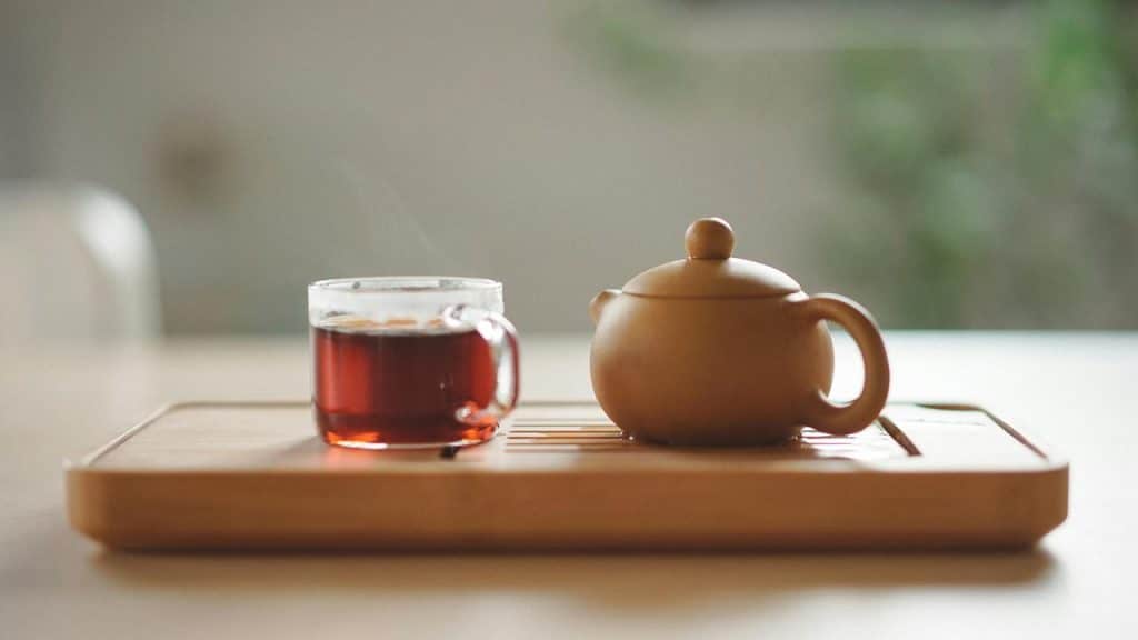 A glass cup of tea and a small clay teapot on a wooden tray.
