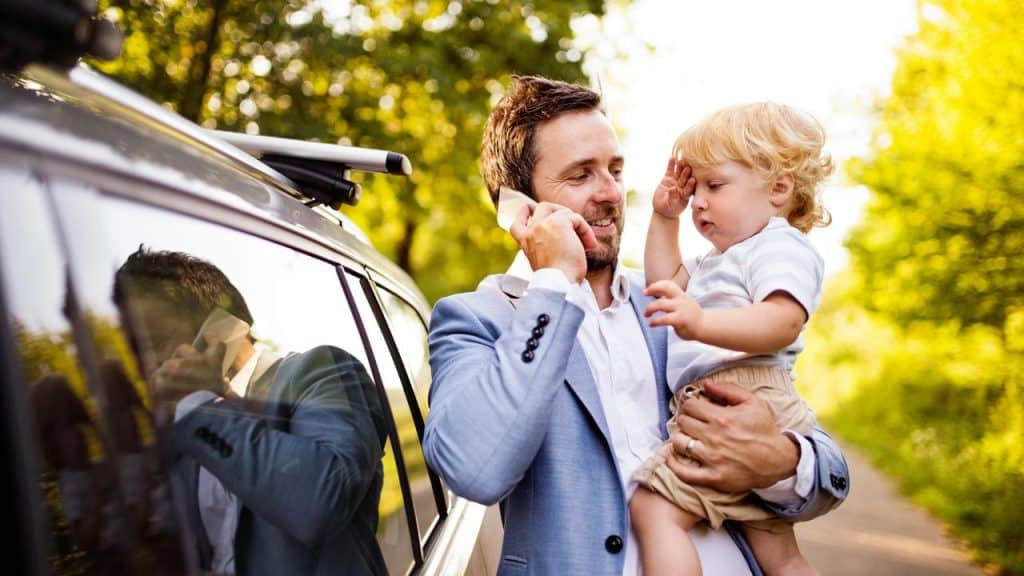 A man holding a toddler and talking on his phone beside a car.