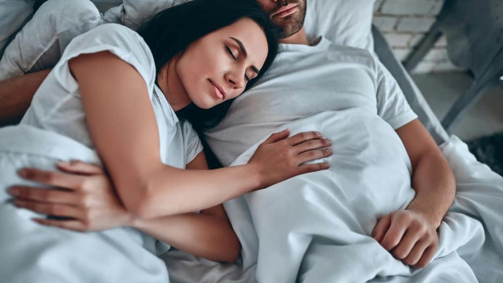 A woman resting her head on her partner’s chest under white bedding.