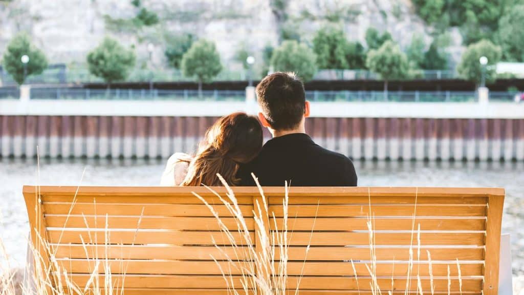 A couple sitting on a bench by the water, one leaning their head on the other's shoulder.