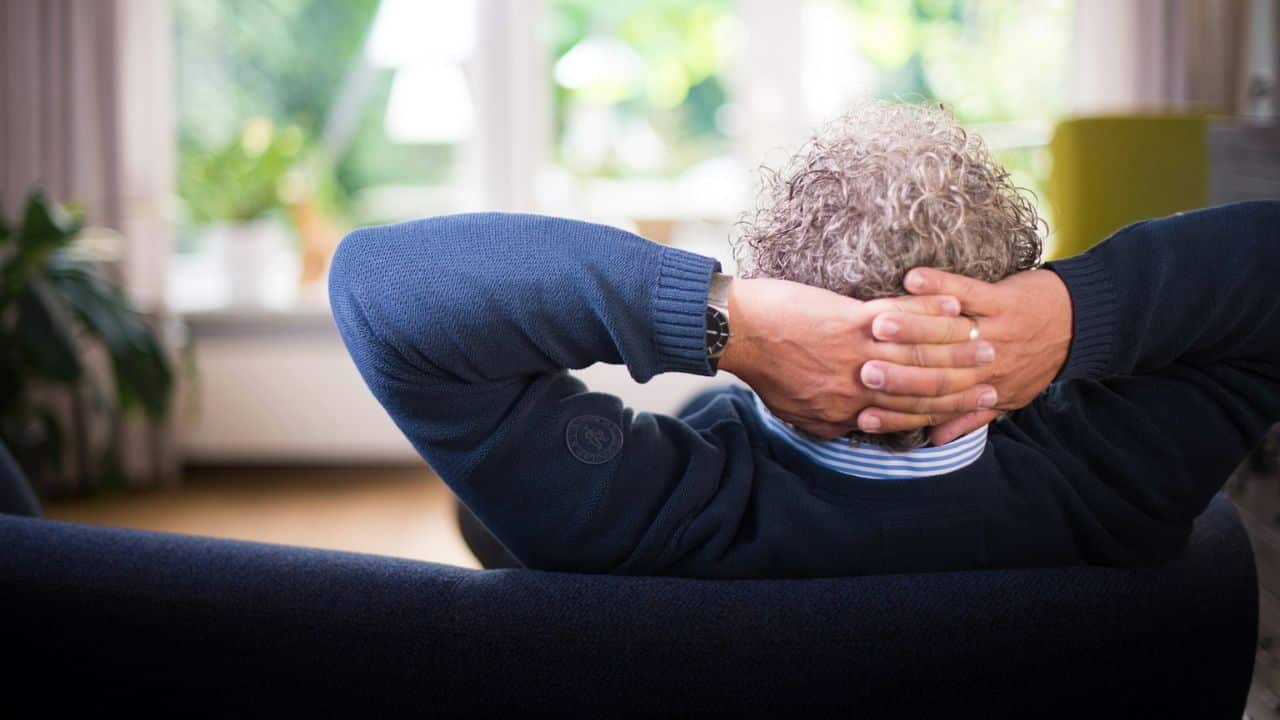 A man with curly grey hair relaxes on a couch, hands behind his head.