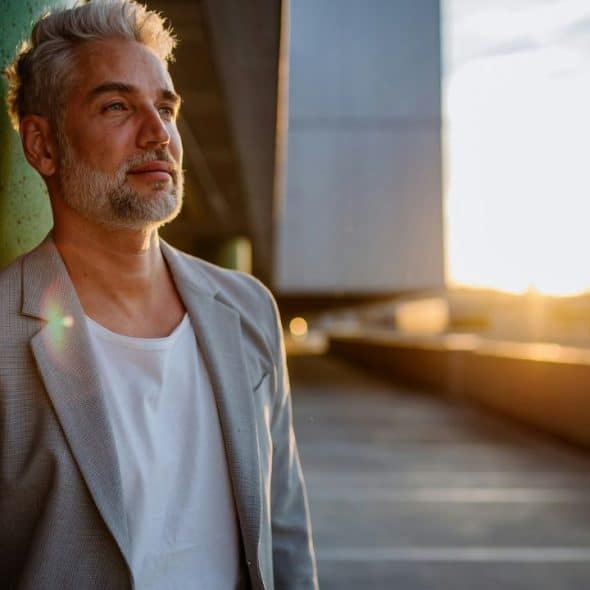 A distinguished man with grey hair and beard, wearing a grey jacket and white t-shirt, gazes pensively into the distance as the sun sets behind him, casting a warm glow.