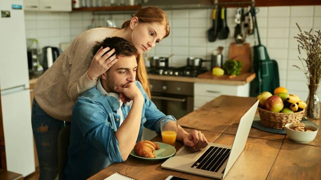 A woman comforts a man working on a laptop at a kitchen table with food and juice.