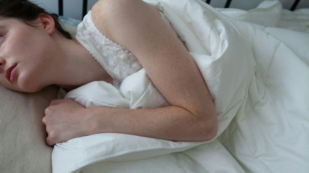A person with freckled arms lies on a bed, covered by a white duvet.