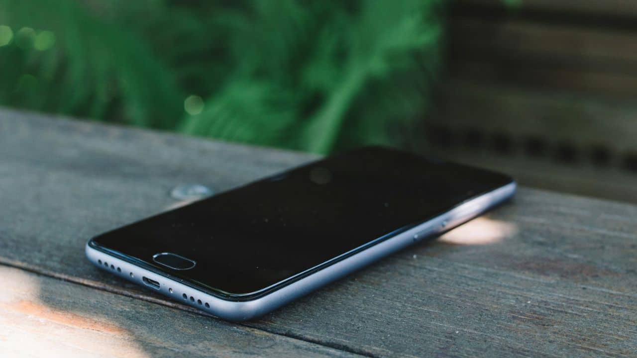 A black smartphone rests on a wooden surface with green foliage in the background.