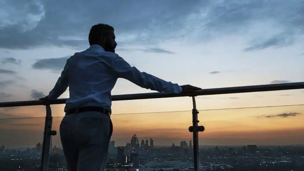 A man in a dress shirt and pants looks out at a city skyline at sunset.