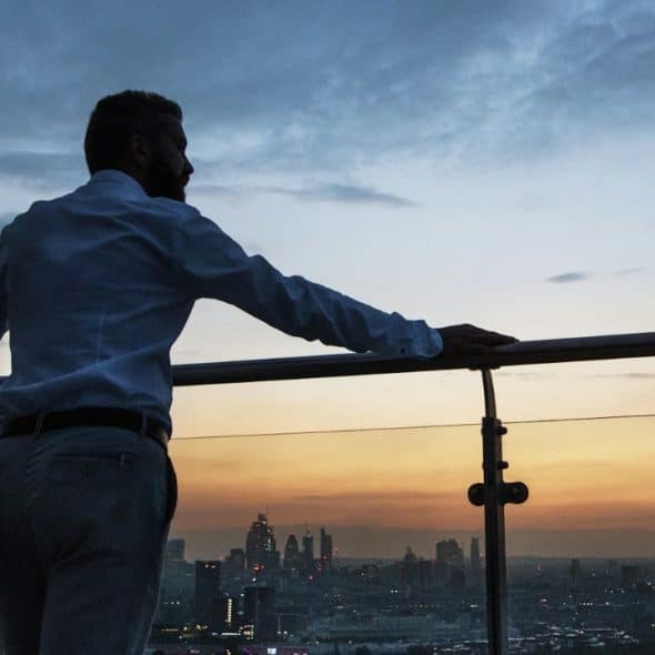 A man in a dress shirt and pants looks out at a city skyline at sunset.