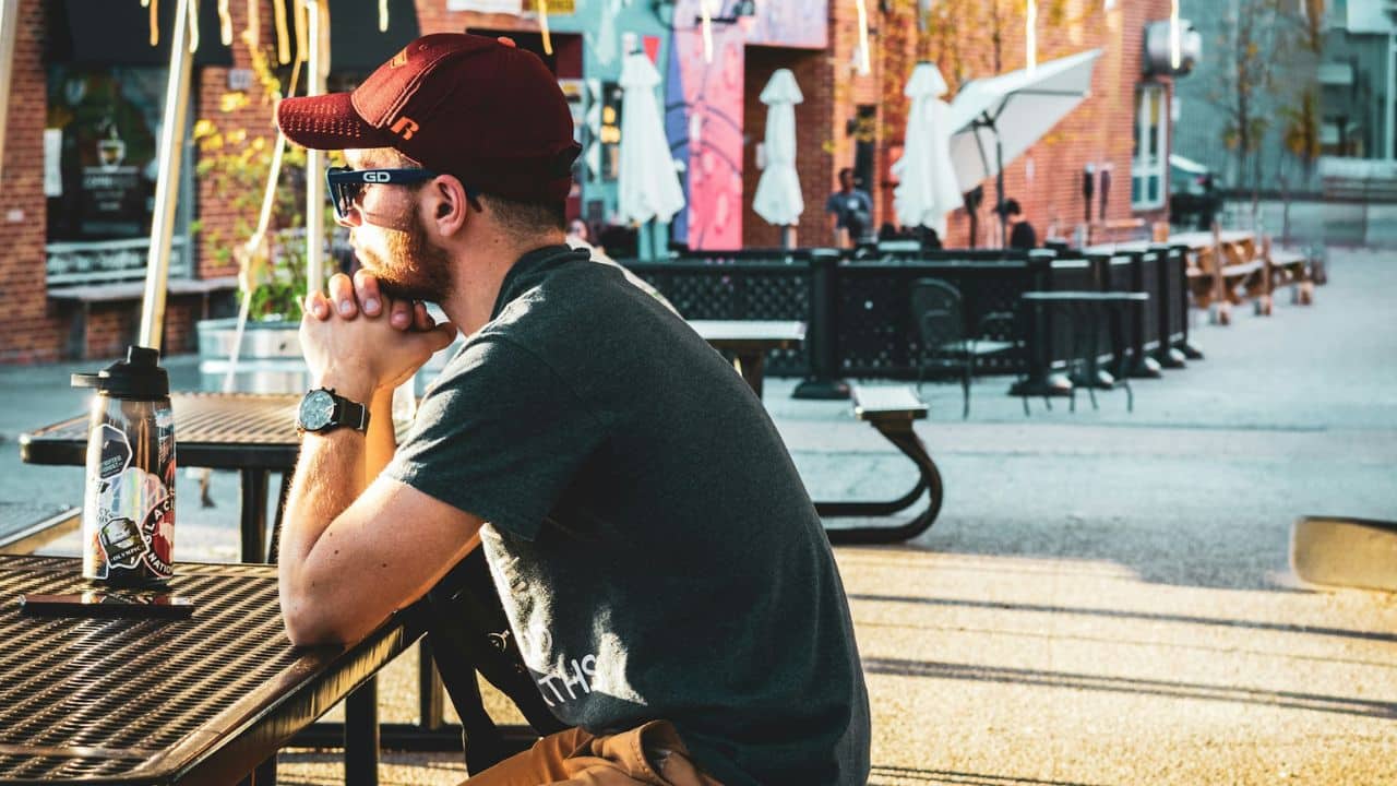 A man in a cap and sunglasses sits at an outdoor table, looking off to the side.