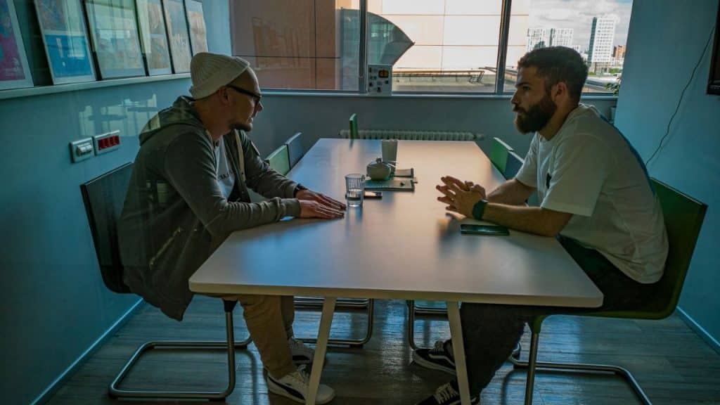 A man in a beanie and glasses sits across a table from a bearded man, both engaged in conversation.