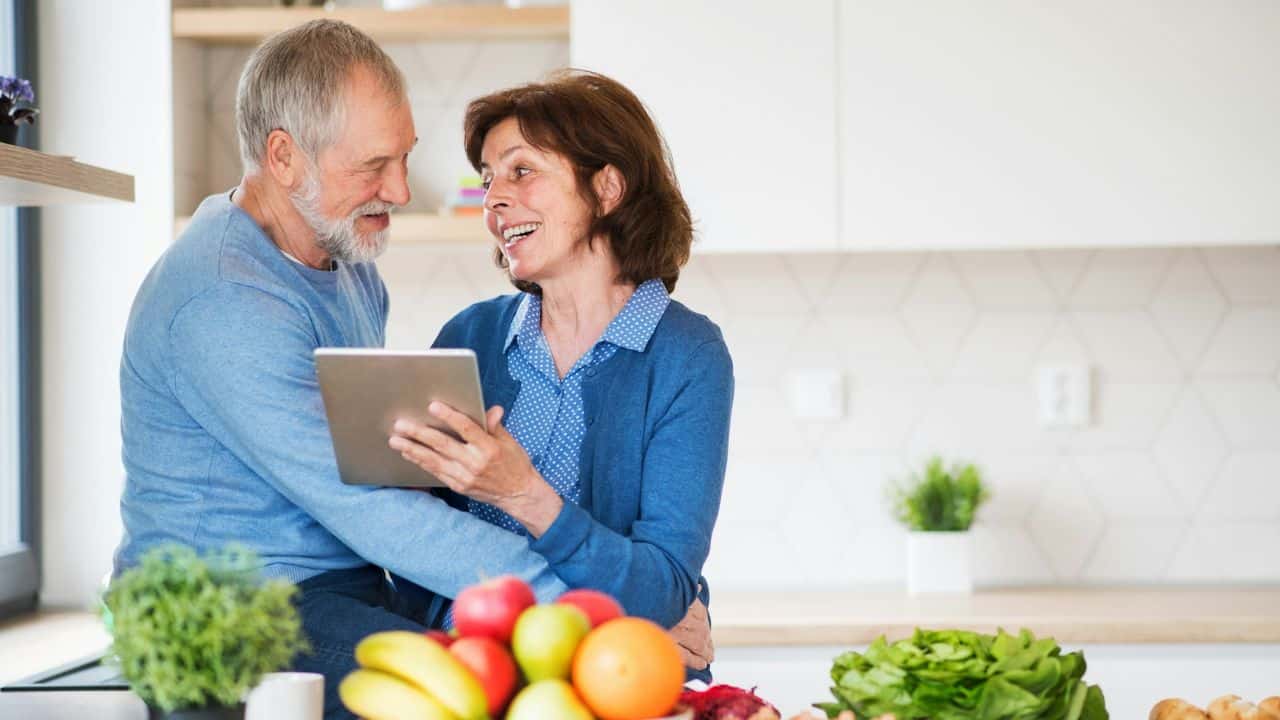 A happy older couple embraces in a kitchen, looking at a tablet together.