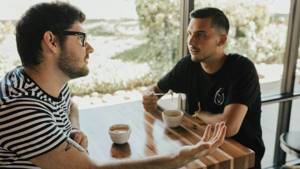 A man in glasses and a striped shirt talks to another man at a table with coffee cups.