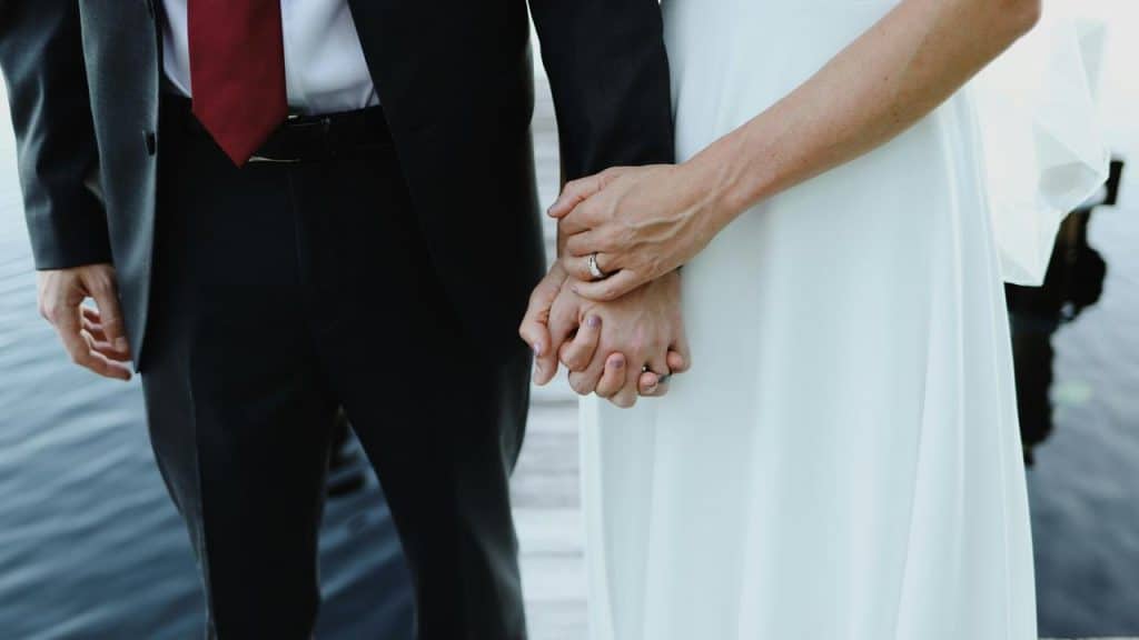 A bride in a white dress and a groom in a dark suit holding hands on a dock over water.