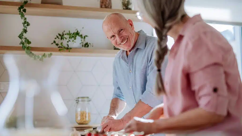 A smiling older man chopping vegetables with his partner in a sunlit kitchen.