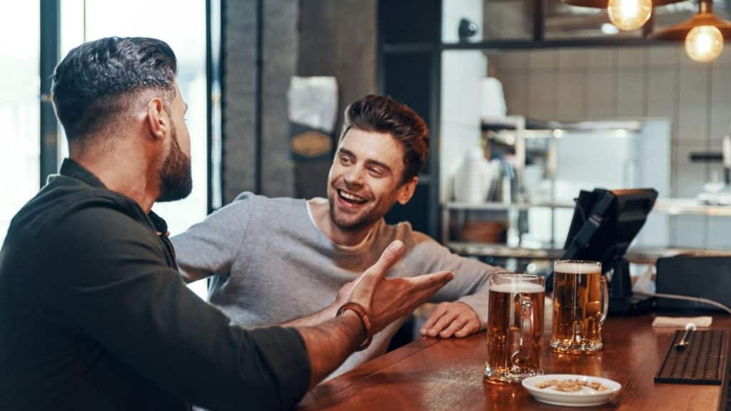 A smiling man with light brown hair sits at a bar, talking to another man.
