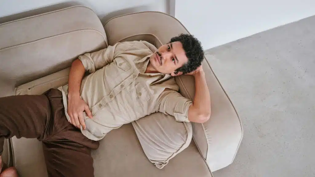 A man with curly hair and a mustache lies on a beige couch, looking up.
