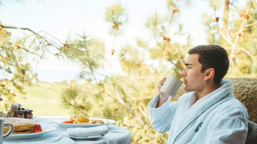 A man in a bathrobe sipping coffee at an outdoor breakfast table.