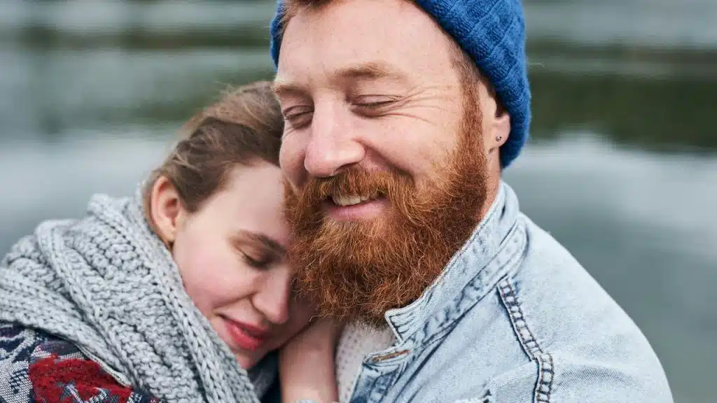 A bearded man in a blue beanie hugging a woman by the water.