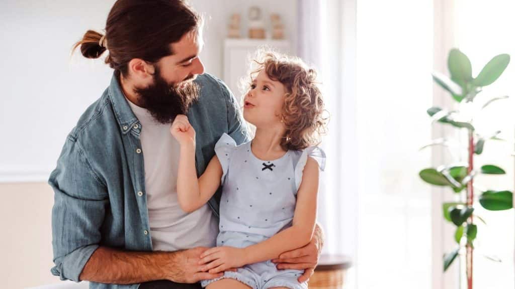 A bearded man holding his daughter on his lap, smiling at her in a sunlit room.