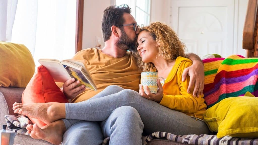 A bearded man kisses a smiling woman's forehead as they relax together on a couch.