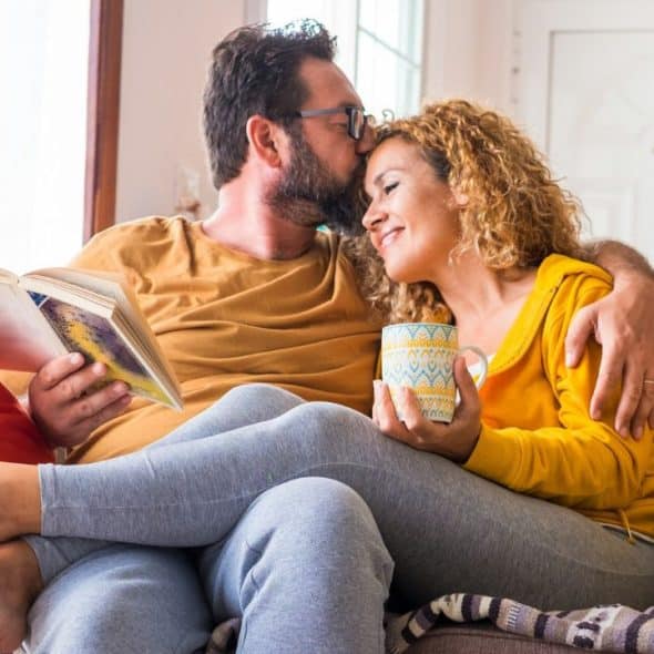 A bearded man kisses a smiling woman's forehead as they relax together on a couch.