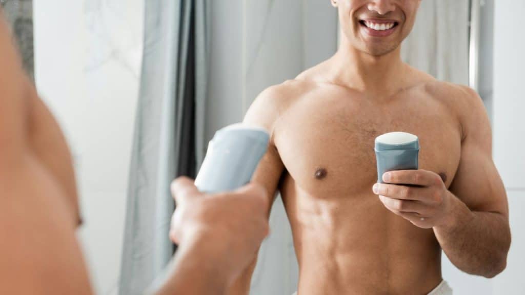 A muscular, smiling man holds up a stick of deodorant while looking in a mirror.