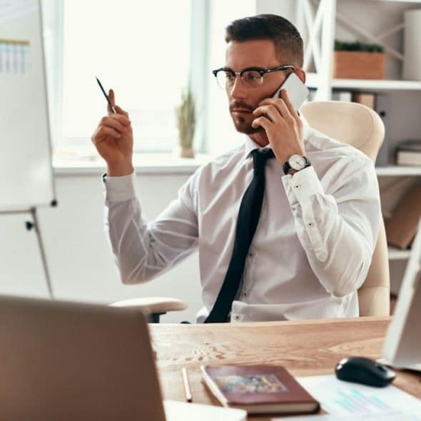 A businessman on the phone holds a pen, looking at a whiteboard.