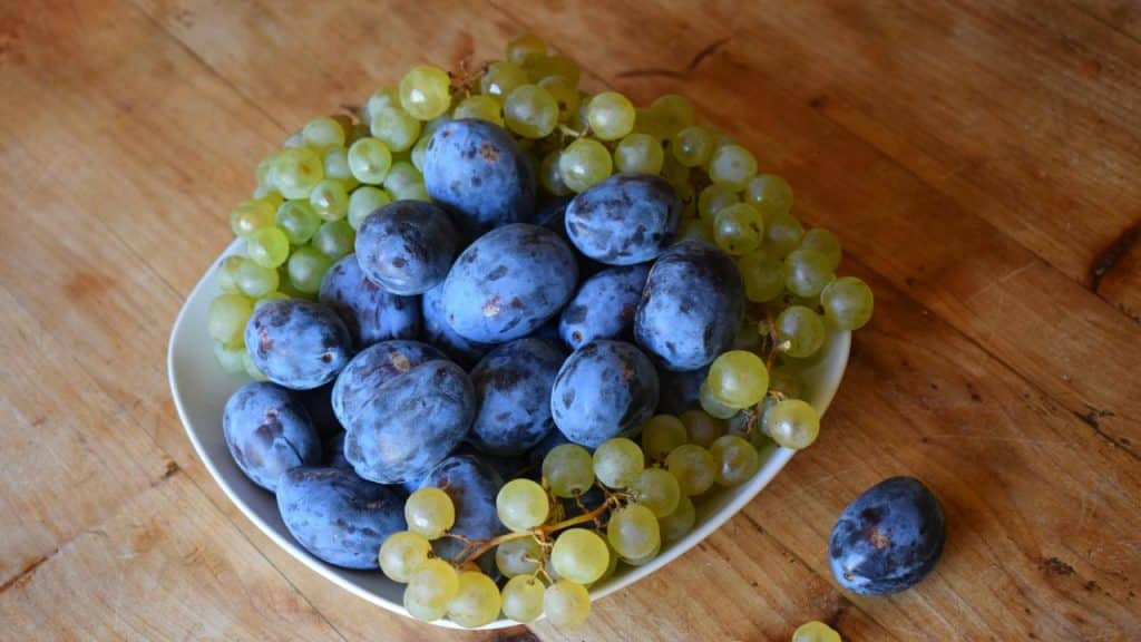 A bowl of blue plums and green grapes on a wooden table.