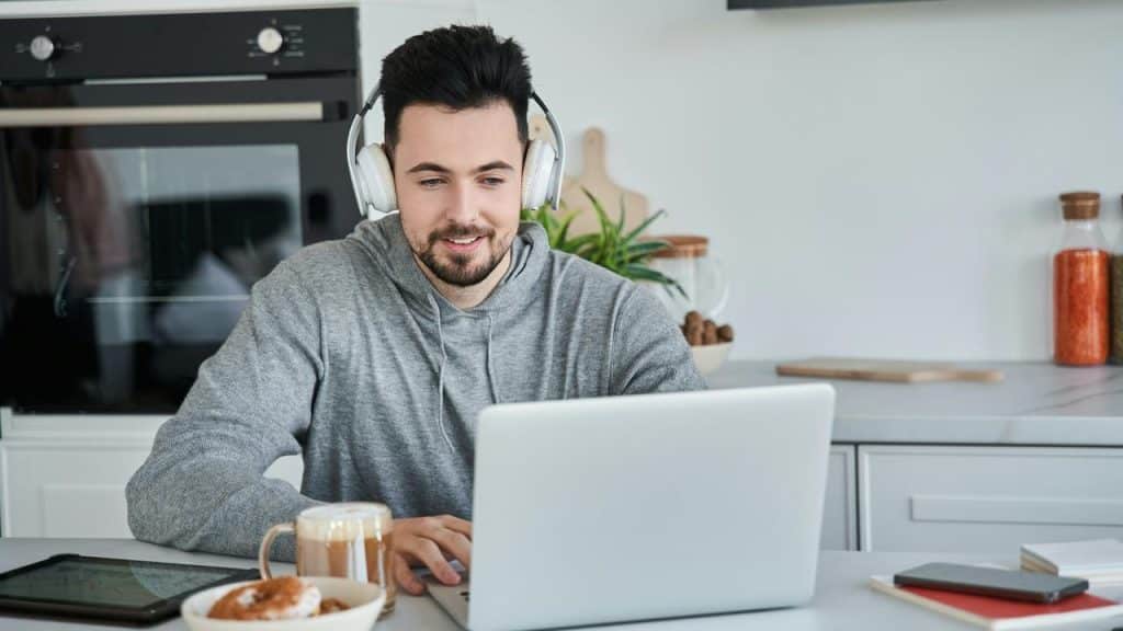 A man wearing headphones working on a laptop at a kitchen counter.