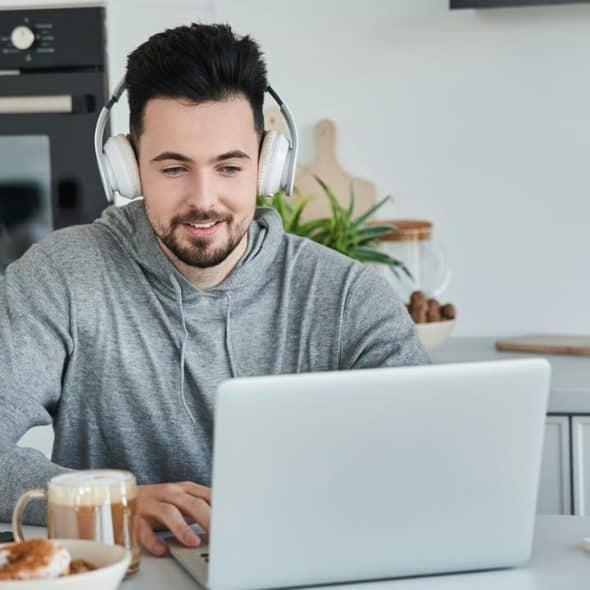 A man wearing headphones working on a laptop at a kitchen counter.