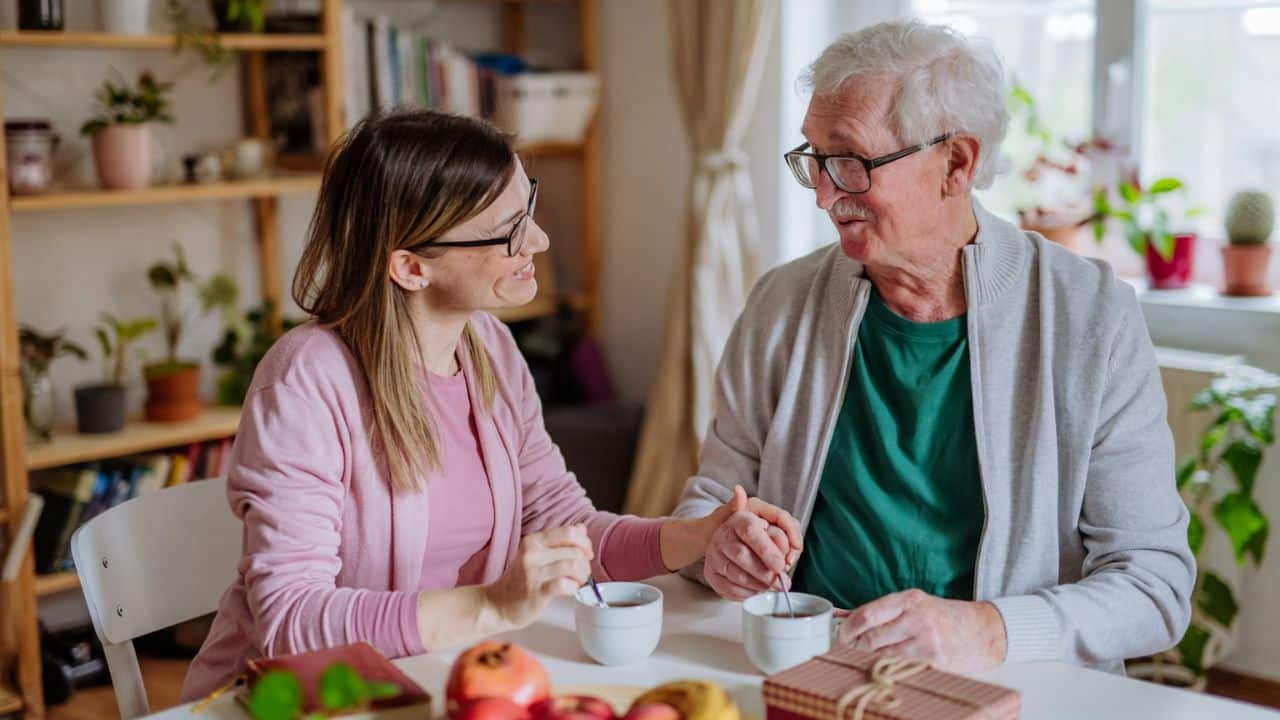 A younger woman and an older man with glasses hold hands at a table, smiling at each other.