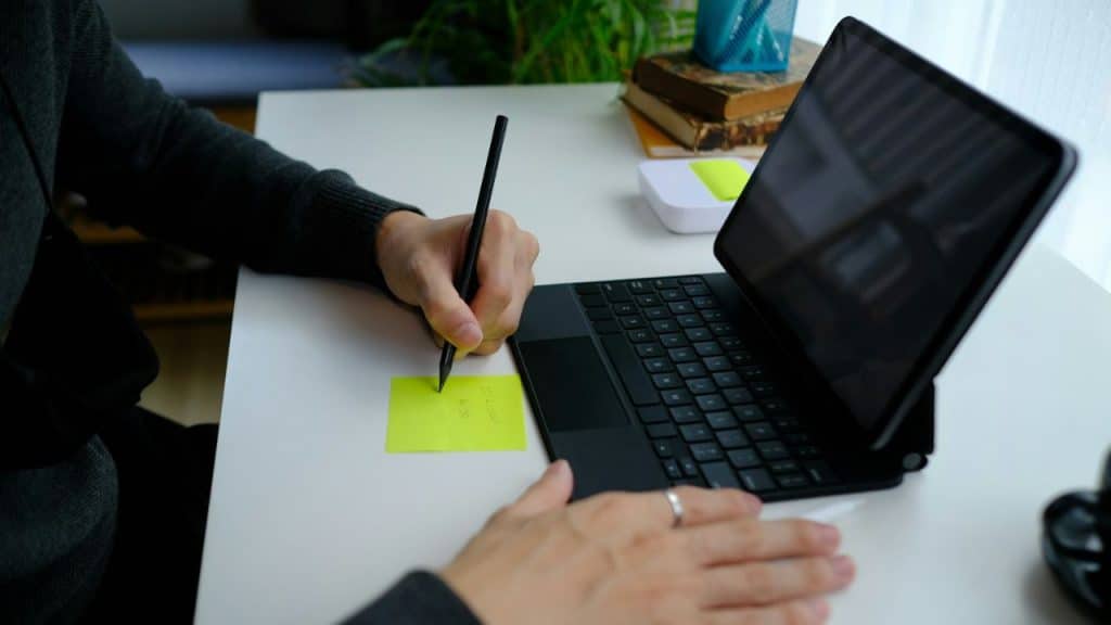 A person's hands are shown writing on a yellow sticky note next to a tablet.