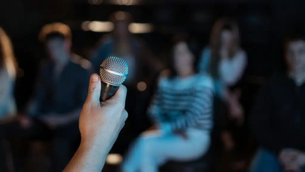 A hand holding a microphone toward a blurred audience.
