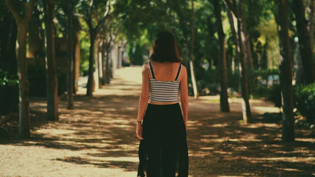 A woman with long dark hair, wearing a striped crop top and black skirt, walks away down a tree-lined path.
