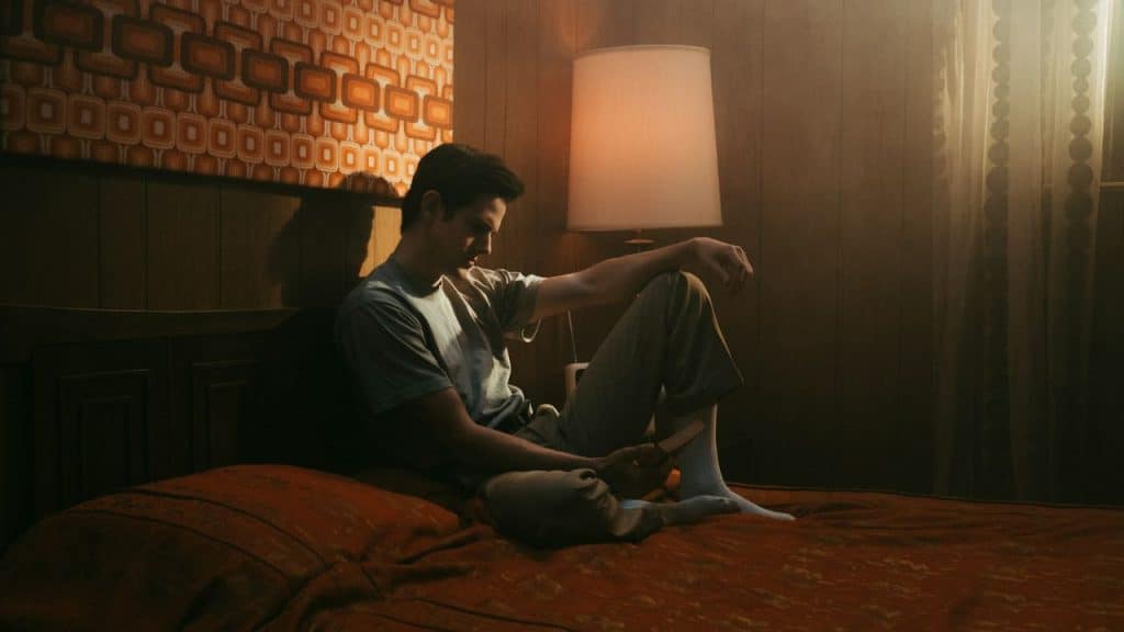 A man sits somberly on a bed in a dimly lit room with vintage decor.