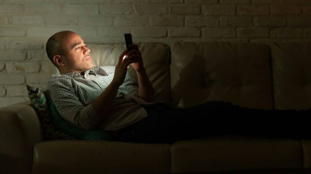 A bald man lies on a couch in a dark room, intently looking at his phone.
