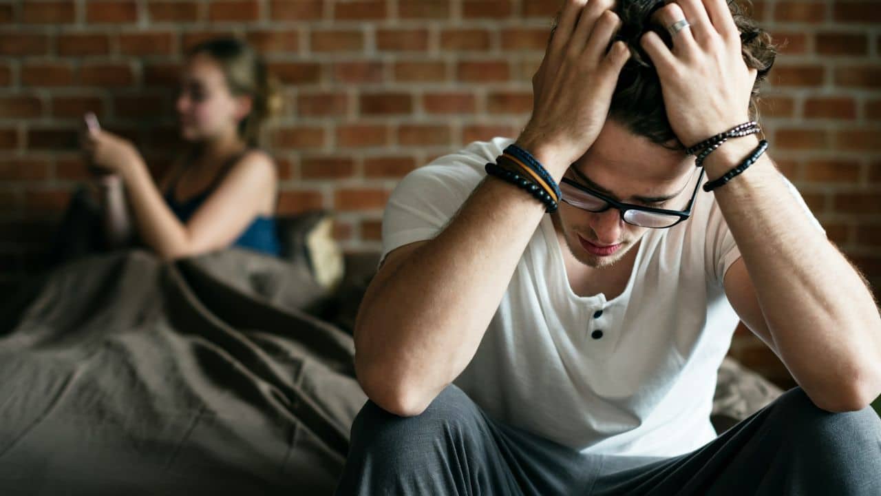 A distressed man with hands in his hair sits in the foreground, with a woman on her phone in the blurry background.