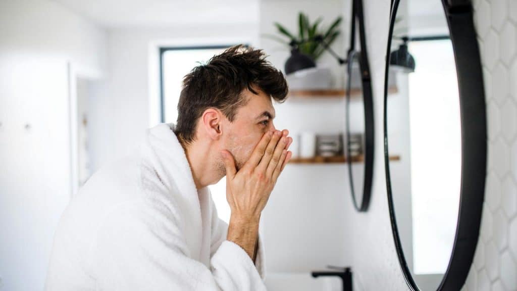 A man in a white robe washing his face at a round bathroom mirror.