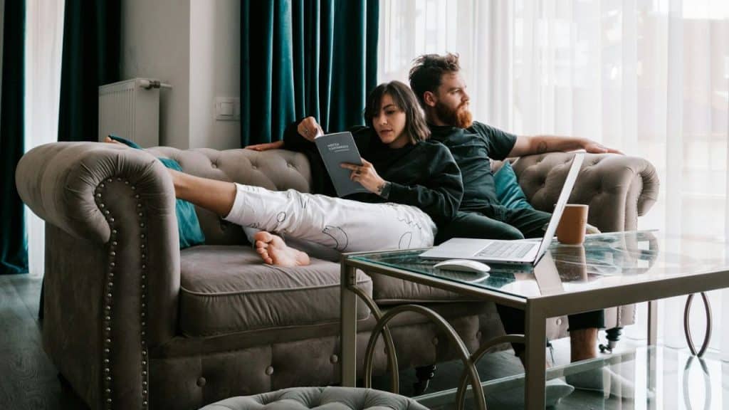 A couple relaxes on a sofa, one reading a book, the other looking away.