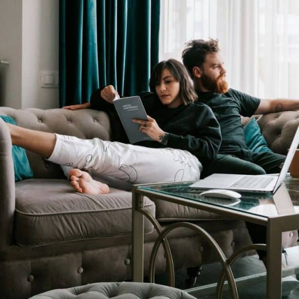 A couple relaxes on a sofa, one reading a book, the other looking away.