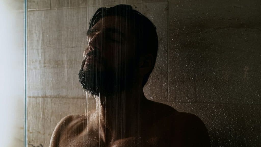 A man with a beard stands in a shower with his eyes closed, water running over his face.