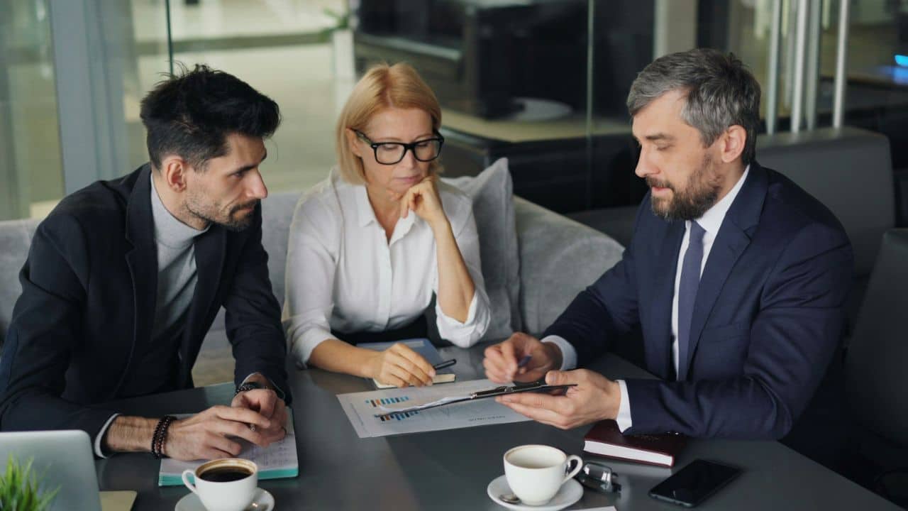 A man in a suit points to a document while two people, a man and a woman, listen intently.
