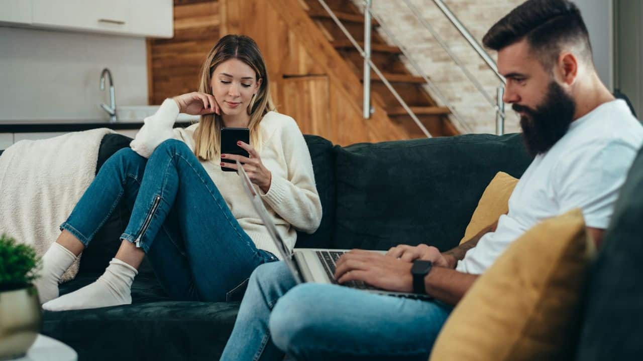 A woman sits on a couch looking at her phone, while a man with a beard sits beside her using a laptop.