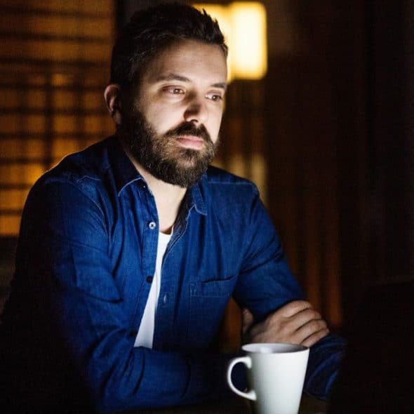 A pensive man with a beard sits in a dimly lit room, a white mug in front of him.