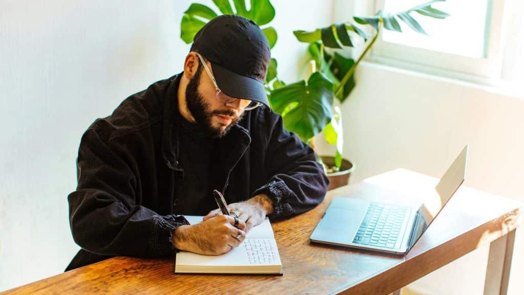 A man writing in a notebook at a desk with a laptop.