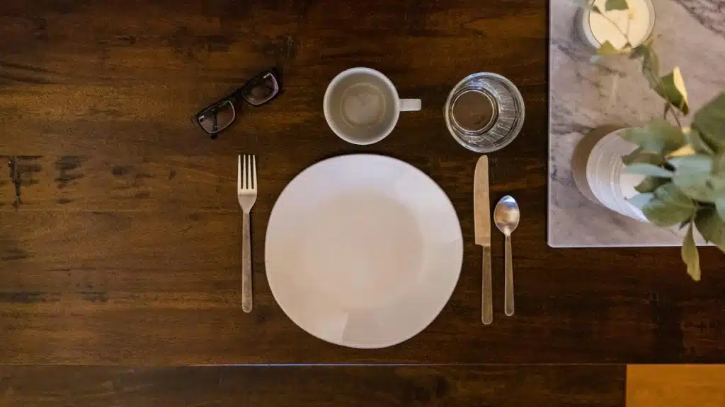 A place setting with a white plate, utensils, mug, and glass on a wooden table.